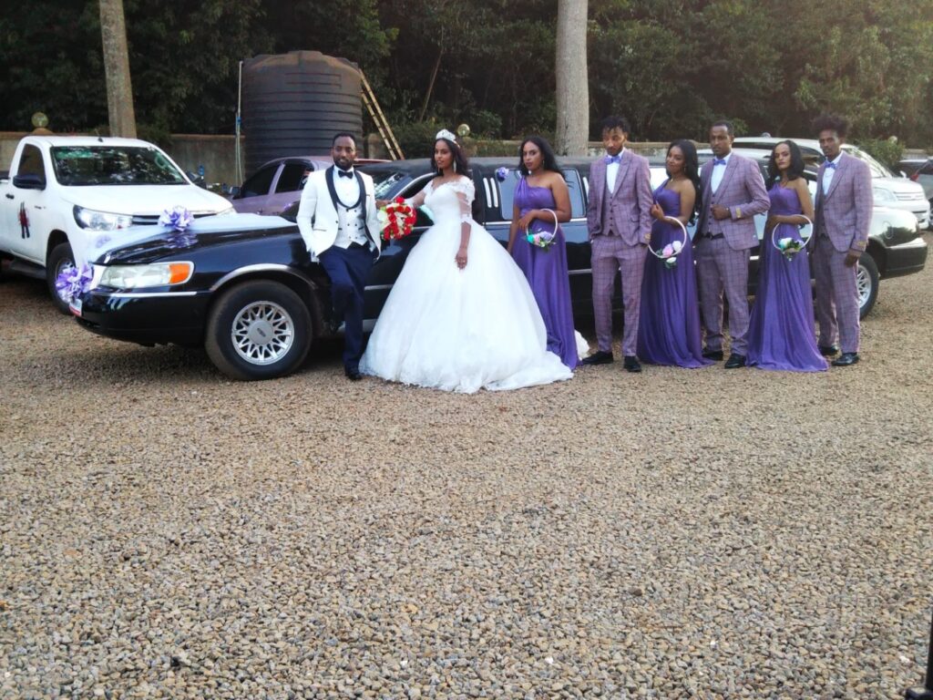 Bride in white ballroom gown and groom in white jacket pose beside a decorated black car in a gravel lot, bridal party in purple gowns and plaid suits nearby with bouquets.