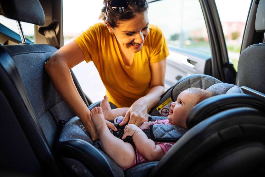 Mother in a yellow shirt adjusts a baby in a car seat inside a car, smiling as she helps.