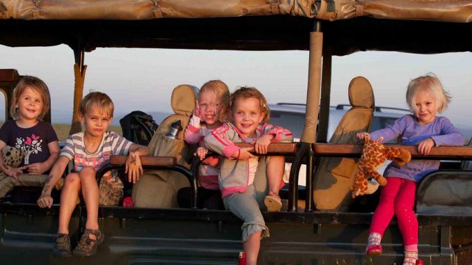 Five young children sit on the open safari vehicle seats at the beach at sunset, smiling and holding stuffed animals.